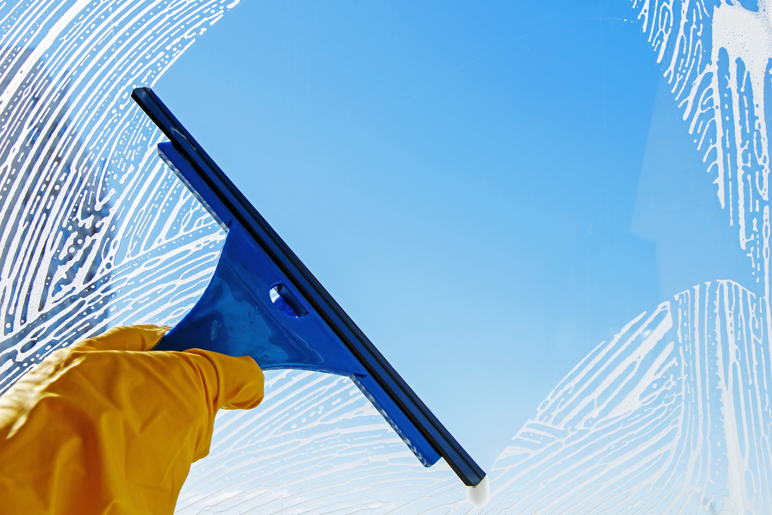 A person with a yellow glove using a squeegee to clean the soap off of a window.